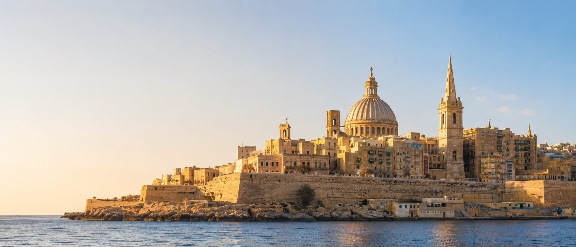 The limestone skyline of Valletta at golden hour — the Carmelite dome and St Paul's spire rising above the fortress walls and the sea